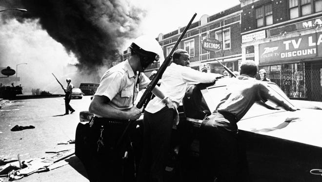 A policeman searches black suspects in a Detroit street on July 25, 1967 as buildings are burning during riots that erupted.