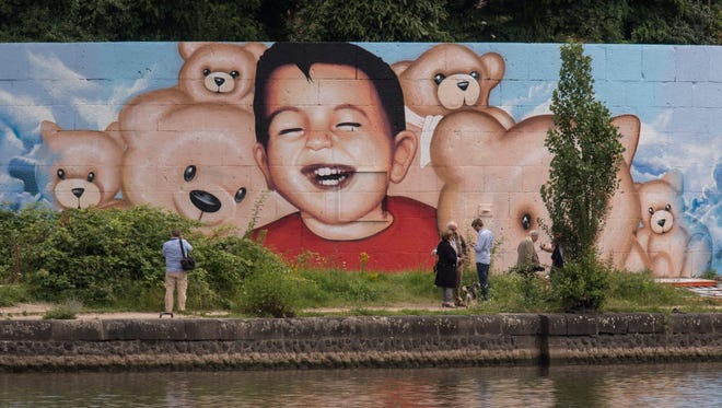 People stand in front of a new mural by artists Justus Becker and Oguz Sen depicting the drowned Syrian refugee boy Alan Kurdi in Frankfurt, Germany, on July 4, 2016. The artists created the mural with the boy surrounded by teddy bears after vandals had destroyed a previous one showing the toddler drowned.